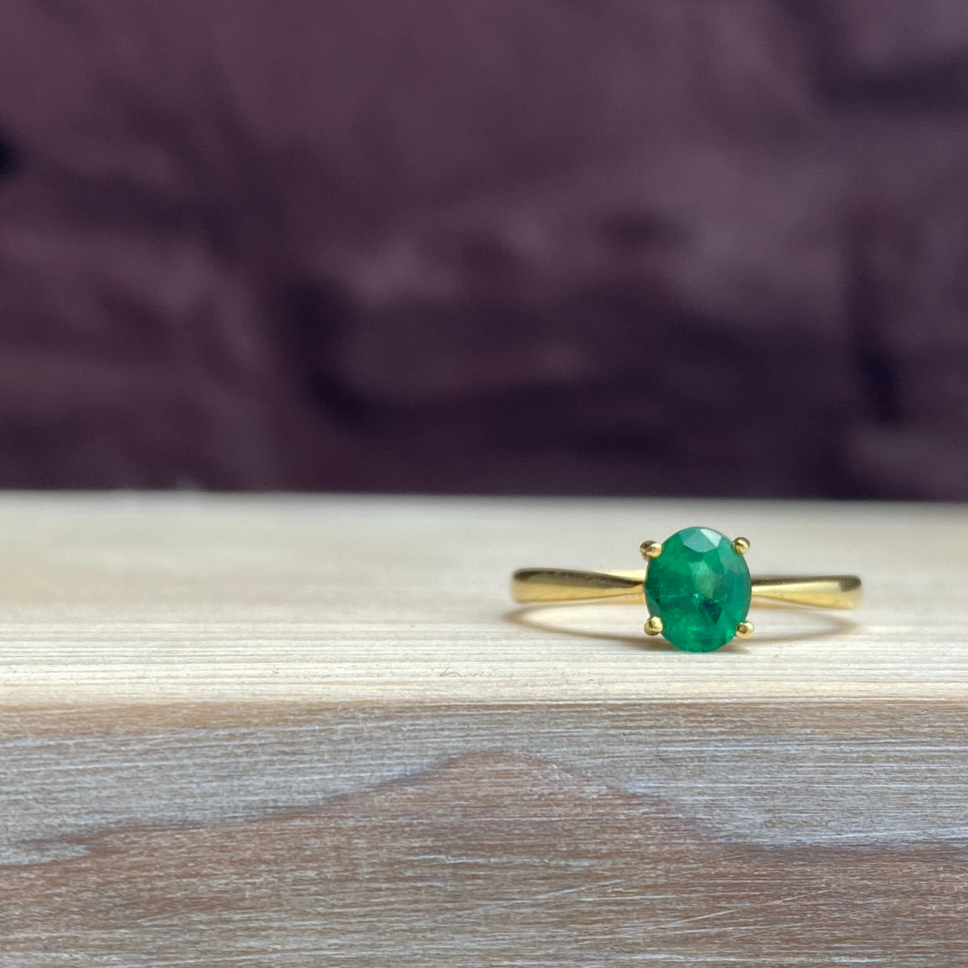 Oval faceted Emerald gemstone with four claw setting in 18ct yellow gold fine ring band on a wooden surface and purple background wall