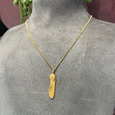 A gold plated sycamore gap seed pendant on a gold curb chain on a grey bust in a jewellery shop in Corbridge 