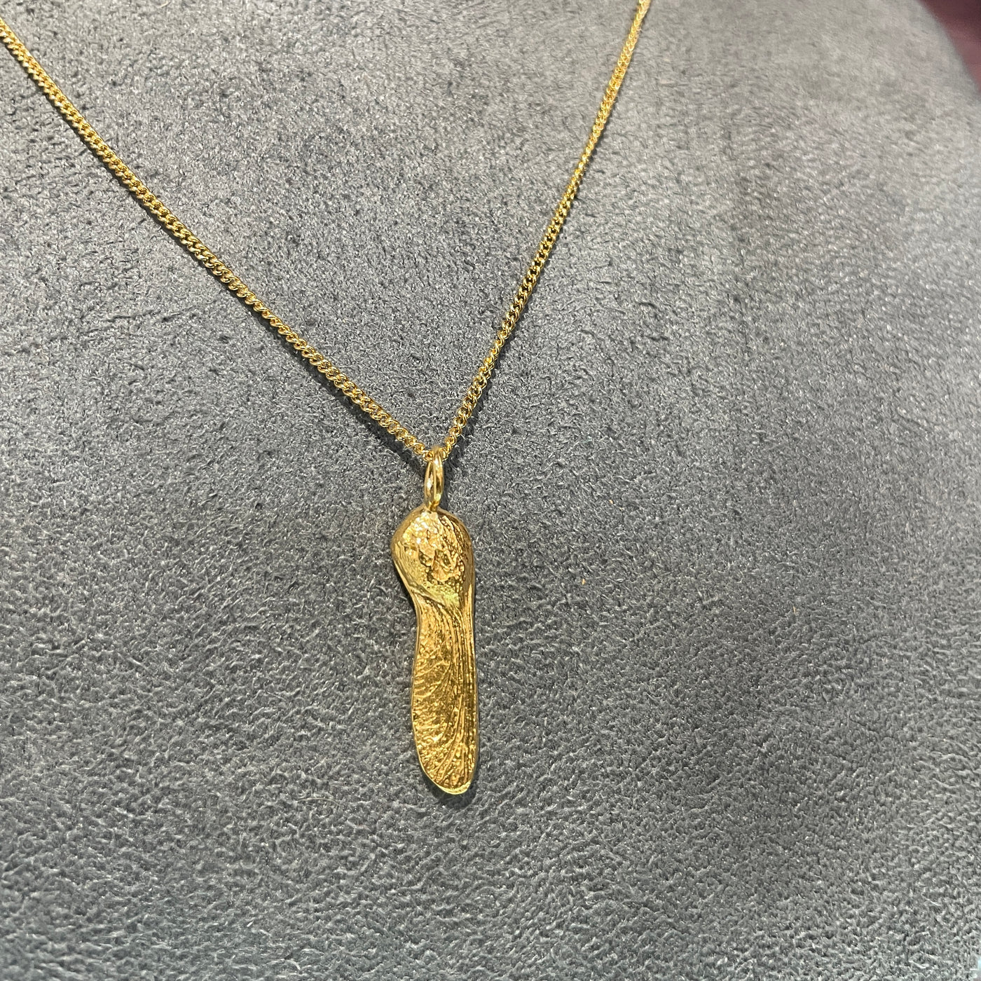 A gold single sycamore seed pendant from Sycamore Gap Ona gold curb chain  on a grey velvet background. 