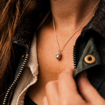 A sterling silver acorn pendant hanging on a silver curb chain from a woman's neck, she is wearing a Barbour jacket which she holds, brown hair is seen in the background.