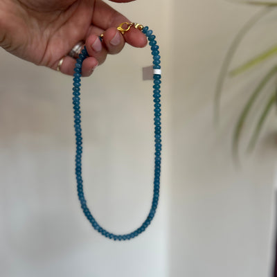 Hand holding a blue beaded necklace with a gold clasp against a neutral background