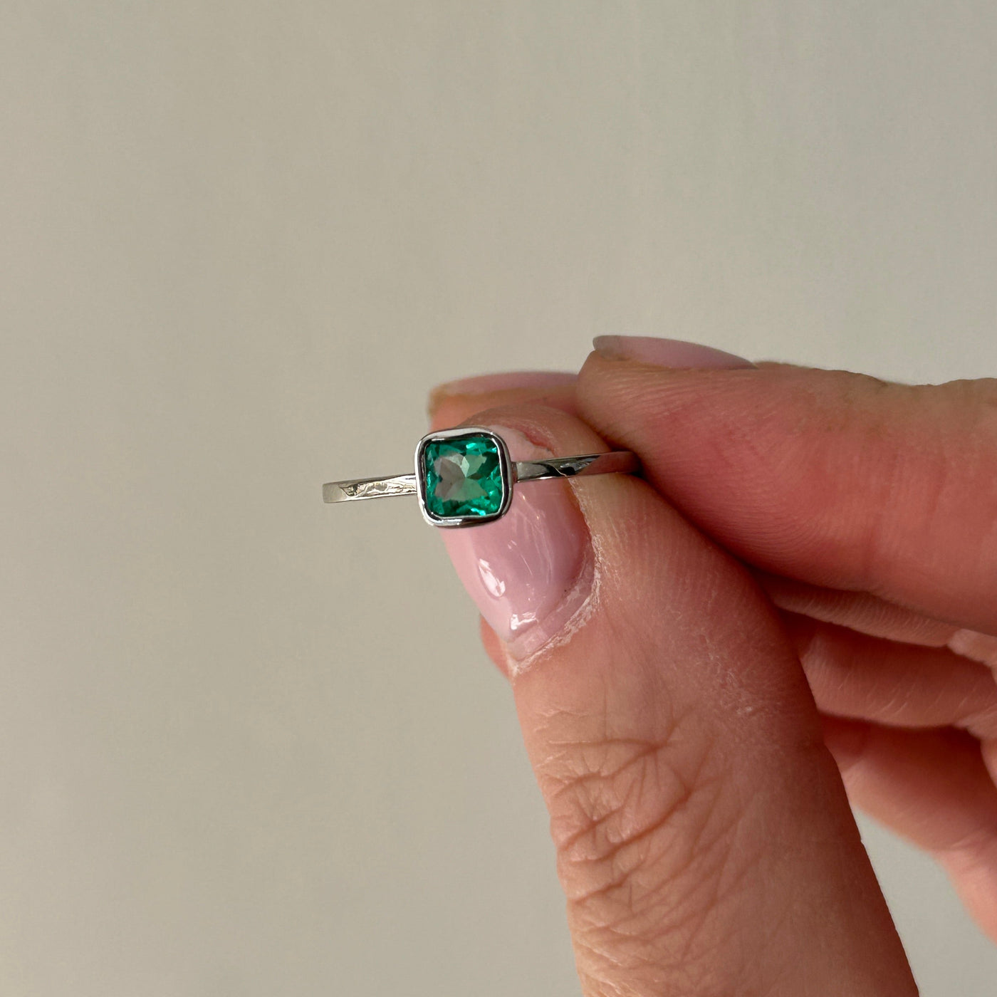 Hand holding a platinum ring with a green emerald gemstone against a neutral background