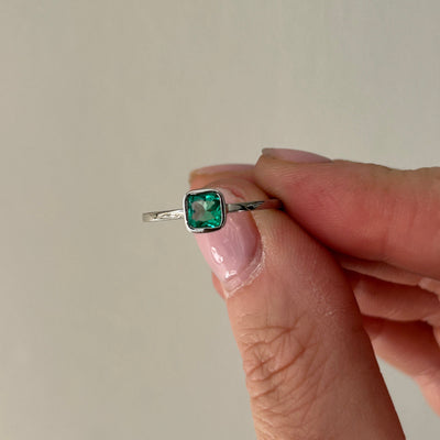 Hand holding a platinum ring with a green emerald gemstone against a neutral background