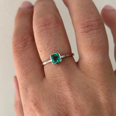 Hand wearing a fine platinum ring with a green emerald gemstone on a neutral background