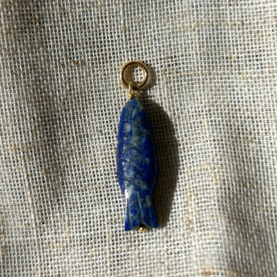 Blue lapis lazuli fish charm with a gold ring on a textured beige background
