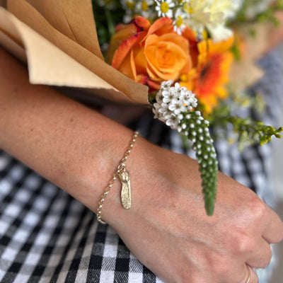 A gold sycamore charm braclelet on a hand holding a bouquet of flowers with a checkered tablecloth in the background