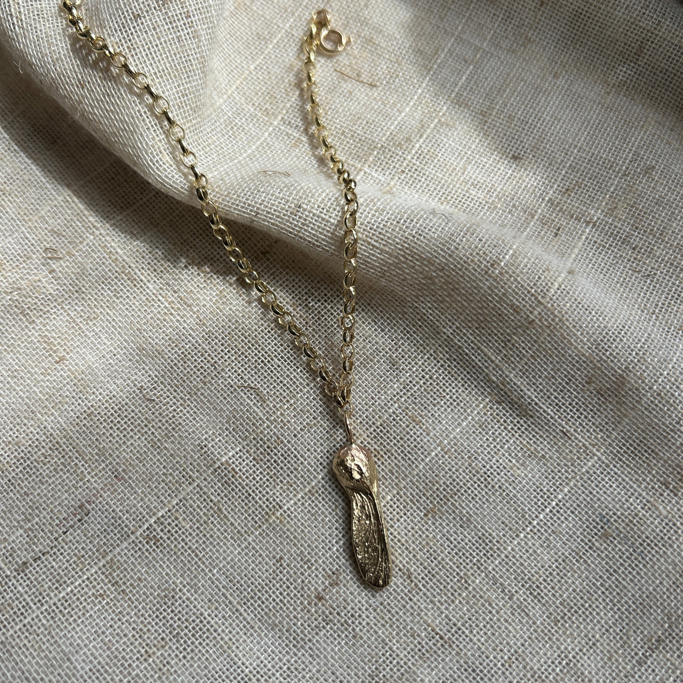 A 9ct yellow gold Sycamore Gap seed charm on a gold oval belcher chain bracelet on a textured linen background.