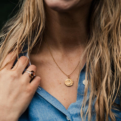 A gold crescent moon and stars gold disk pendant on a gold curb chain worn by a female model wearing a denim shirt and long blonde hair. 