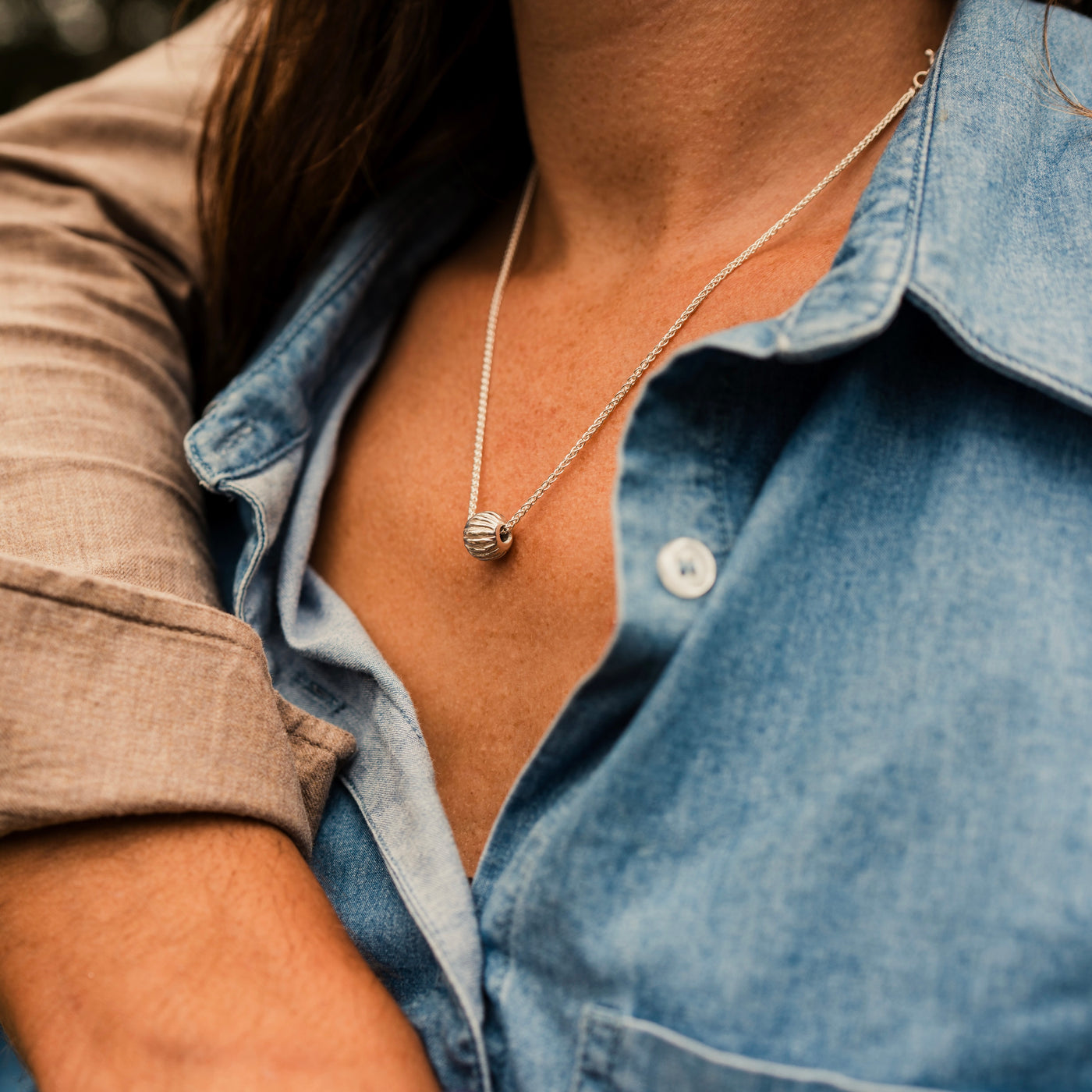 A sterling silver Melon bead threaded onto a silver chunky spiga chain worn by a woman wearing a denim shirt and brown hair, a man's arm in a brown shirt is over her shoulder. Handmade in Corbridge at Kirsty Taylor Goldsmiths