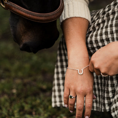 Close-up of a person's hand wearing a bracelet and ring, standing next to a horse in a grassy area.