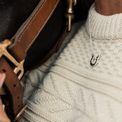 Close-up of a brown leather strap with gold buckle on a dark background, the women is wearing a cream jumper and silver horseshoe pendant