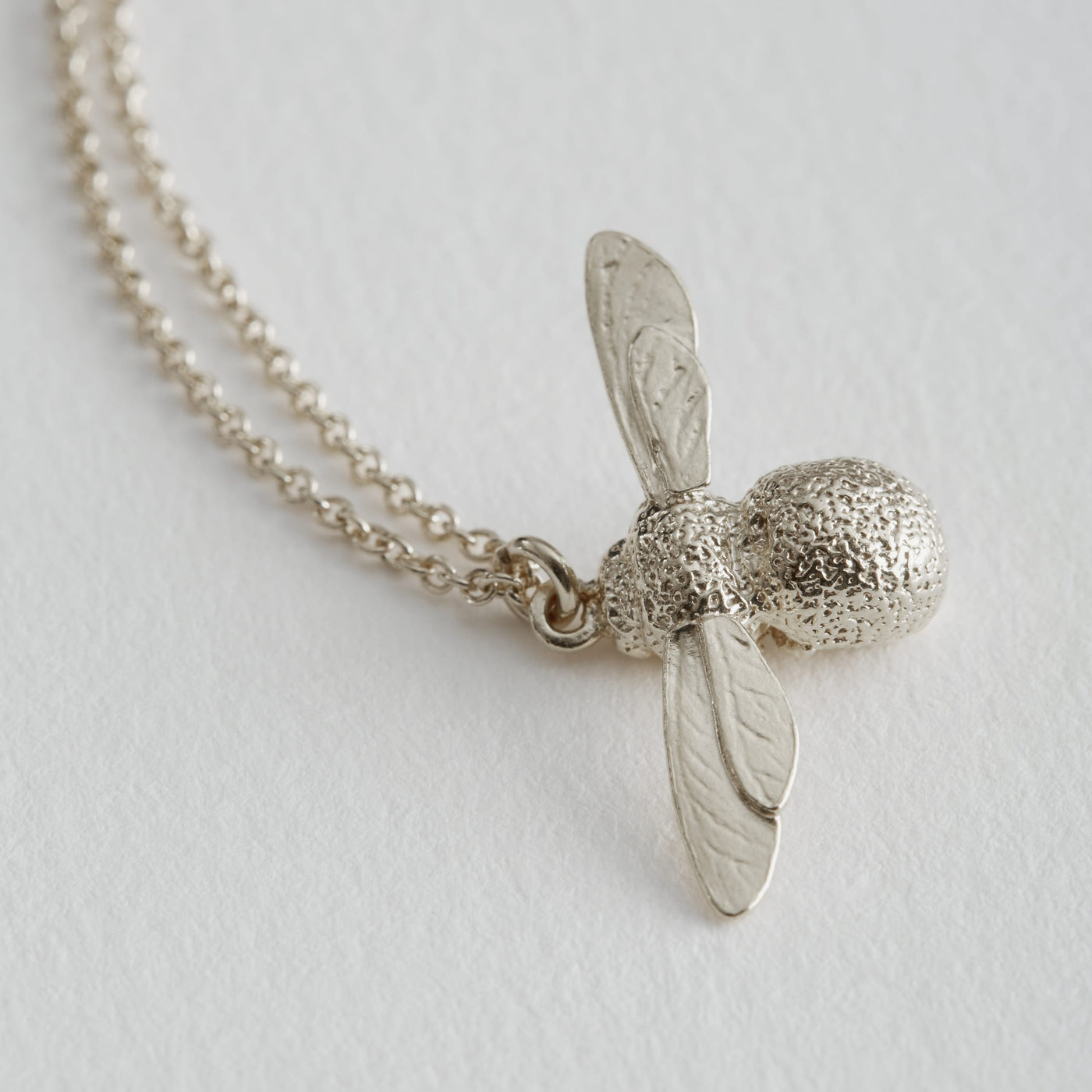 A silver model of a small Bee with outstretched wings with a silver trace chain lying on a white background, made by Alex Monroe and stocked by Kirsty Taylor Jewellery in Northumberland.