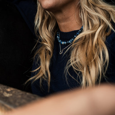 Person with long blonde hair wearing a dark blue sweater and necklace, sitting at a wooden table.