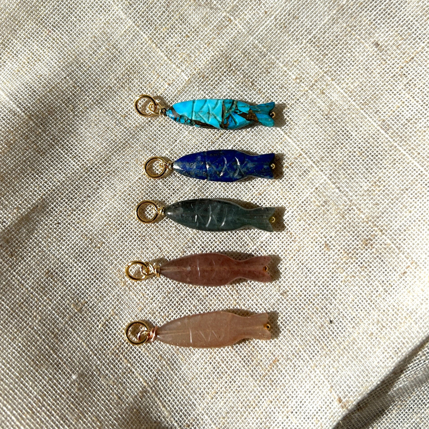 Five small gemstone fish pendants on a textured fabric background