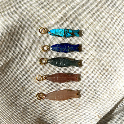 Five small gemstone fish pendants on a textured fabric background