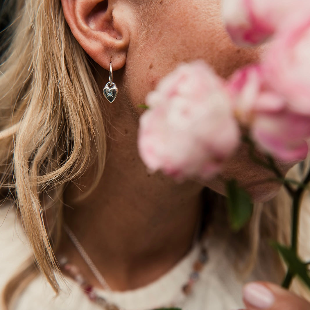 Close-up of a person wearing a heart-shaped earring with pink flowers in the foreground.
