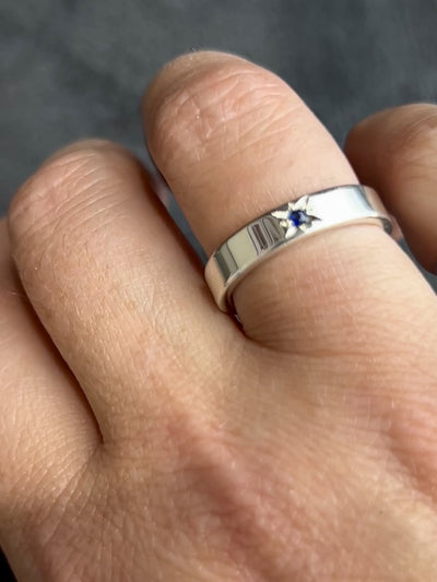 a sterling silver flat ring with a blue sapphire star set in the middle on a hand against a grey background.