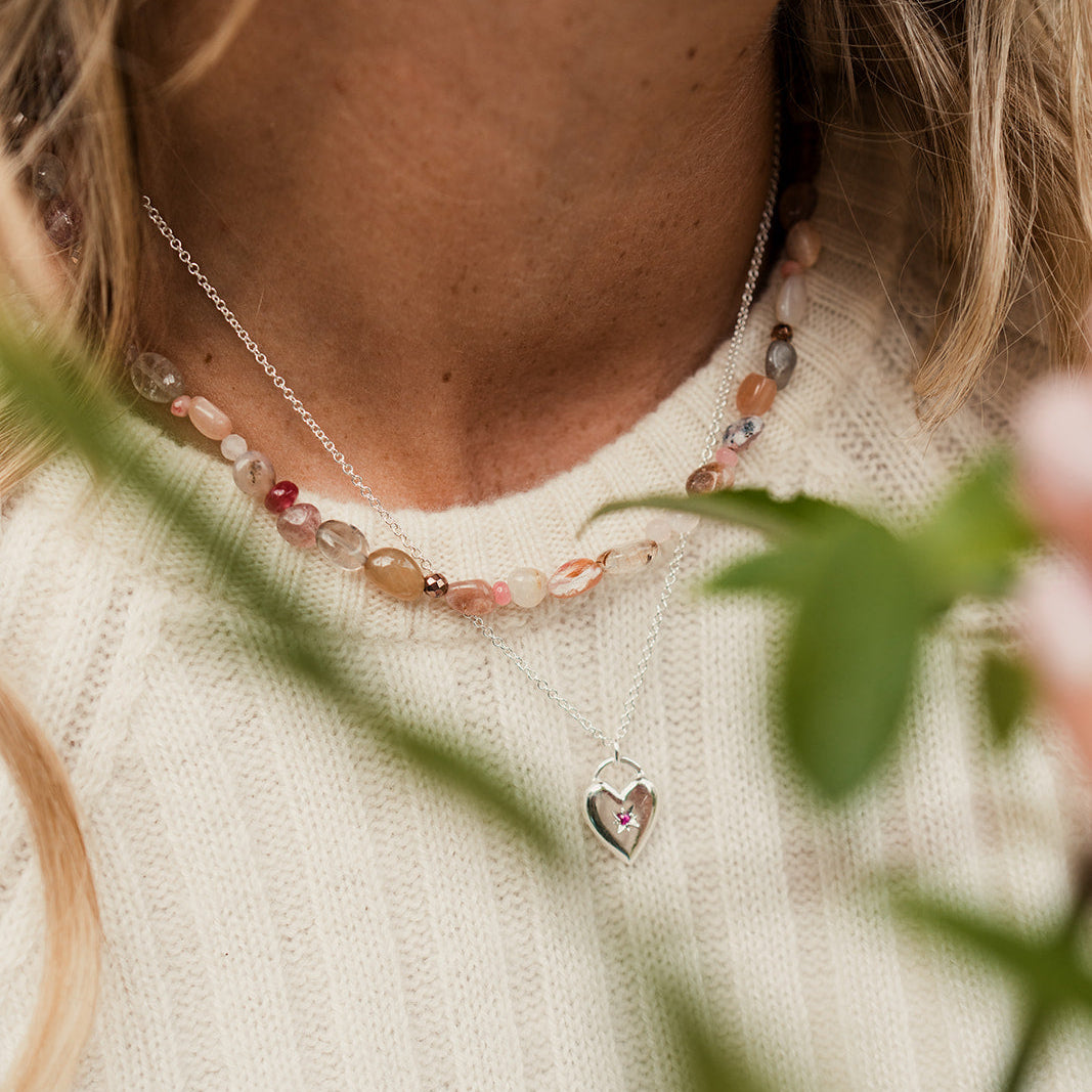 Close-up of a person wearing a necklace with a heart pendant, surrounded by green leaves and flowers.