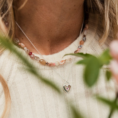 Close-up of a person wearing a necklace with a heart pendant, surrounded by green leaves and flowers.