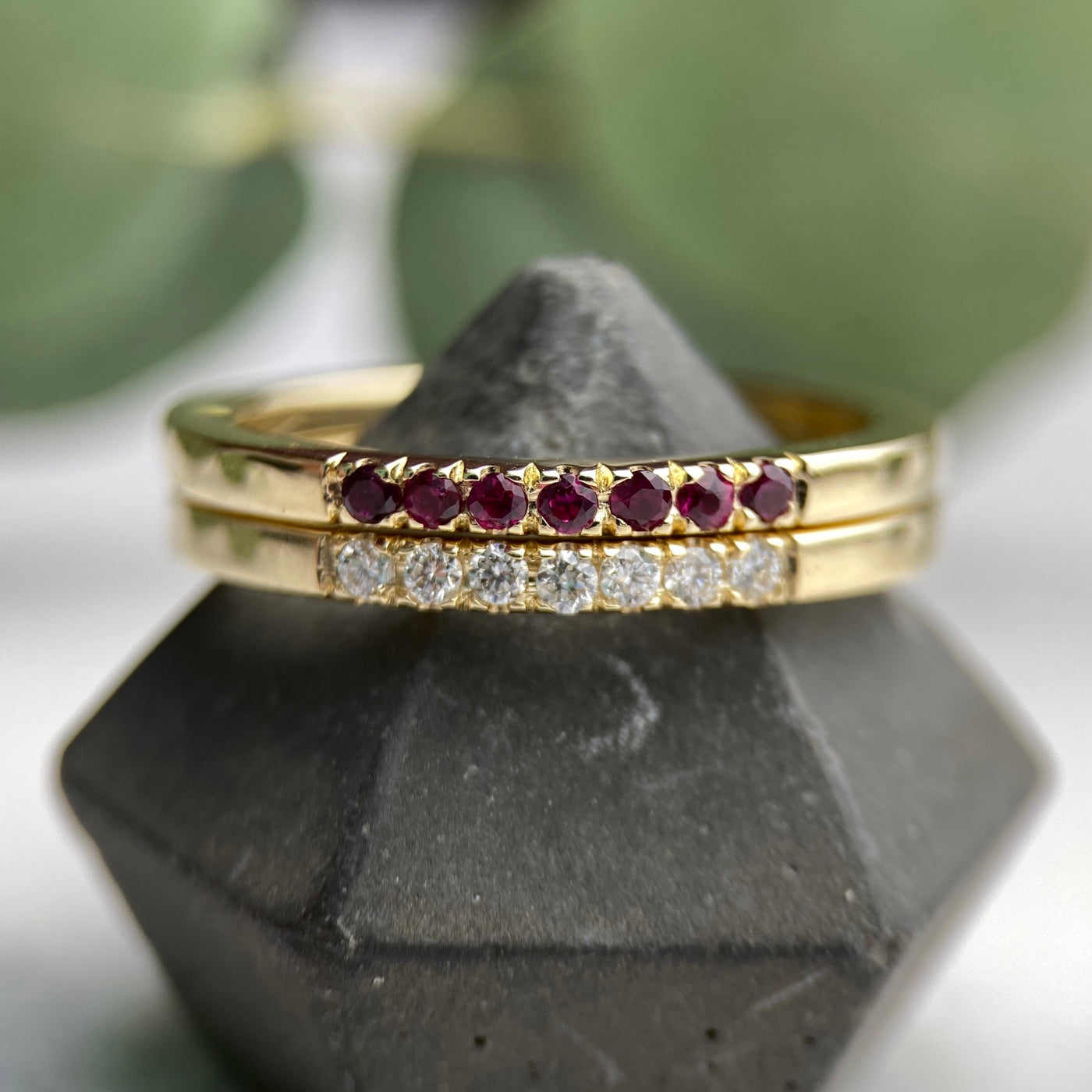 a yellow gold stacking ring measuring 2mm set with 7 round faceted ruby stones pave set into the surface of the ring, the ring is stacked on top another ring set with 7 pave set diamonds as a stacking ring set, both rings are on a grey ring stand with a white and dark red background with a few leaves in view