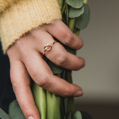 A hand holding a stems of flowers and a yellow cuff of a jumper one finger has a yellow gold love knot proposal ring on the wedding ring finger. Handcrafted gold ring made by Kirsty Taylor Goldsmiths, Corbridge Northumberland