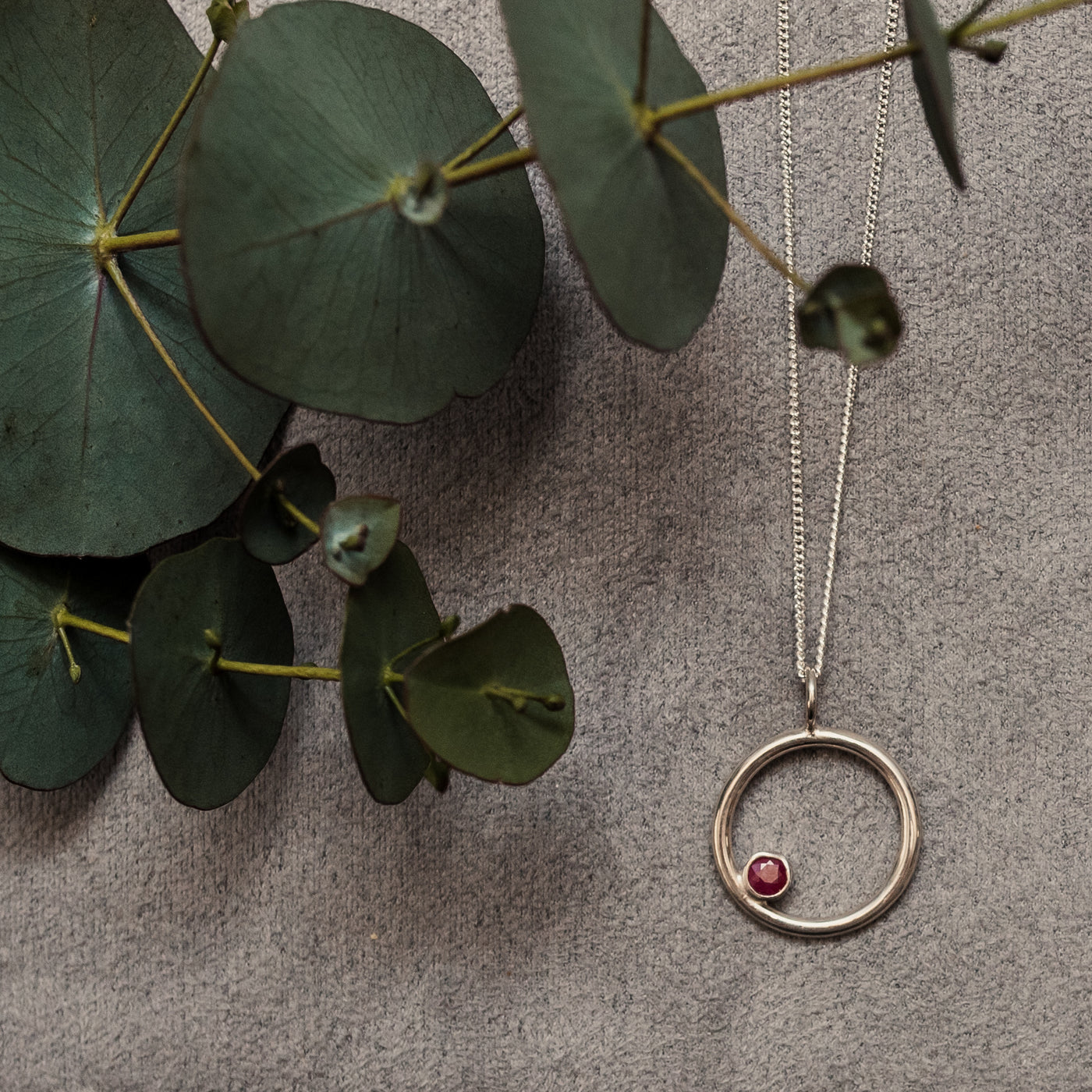 Silver circle pendant with offset bezel set red ruby 4mm round gemstone on a silver chain with a grey velvet background and leaves. Handmade in Corbridge Northumberland