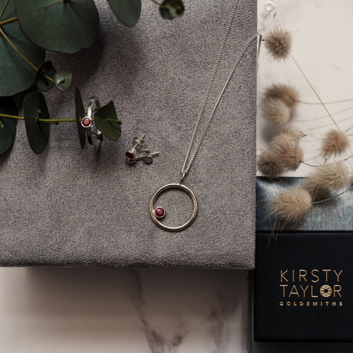 A silver circle necklace with 4mm ruby stone offset on one side on a silver curb chain with a grey velvet background, leaves and a black Kirsty Taylor Jewellery box