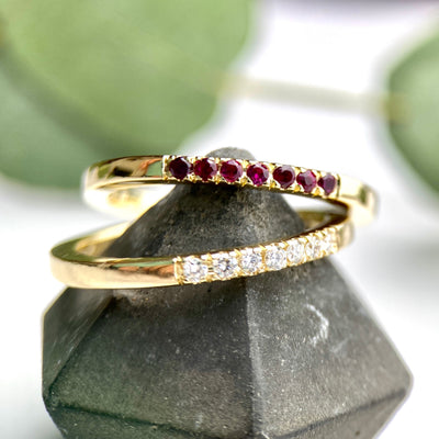 yellow gold stacking ring pave set with 7 round faceted ruby gemstones  placed on top of a yellow gold stacking ring with faceted diamonds on a grey ring block with a white and dark red background and a couple of leaves in the background